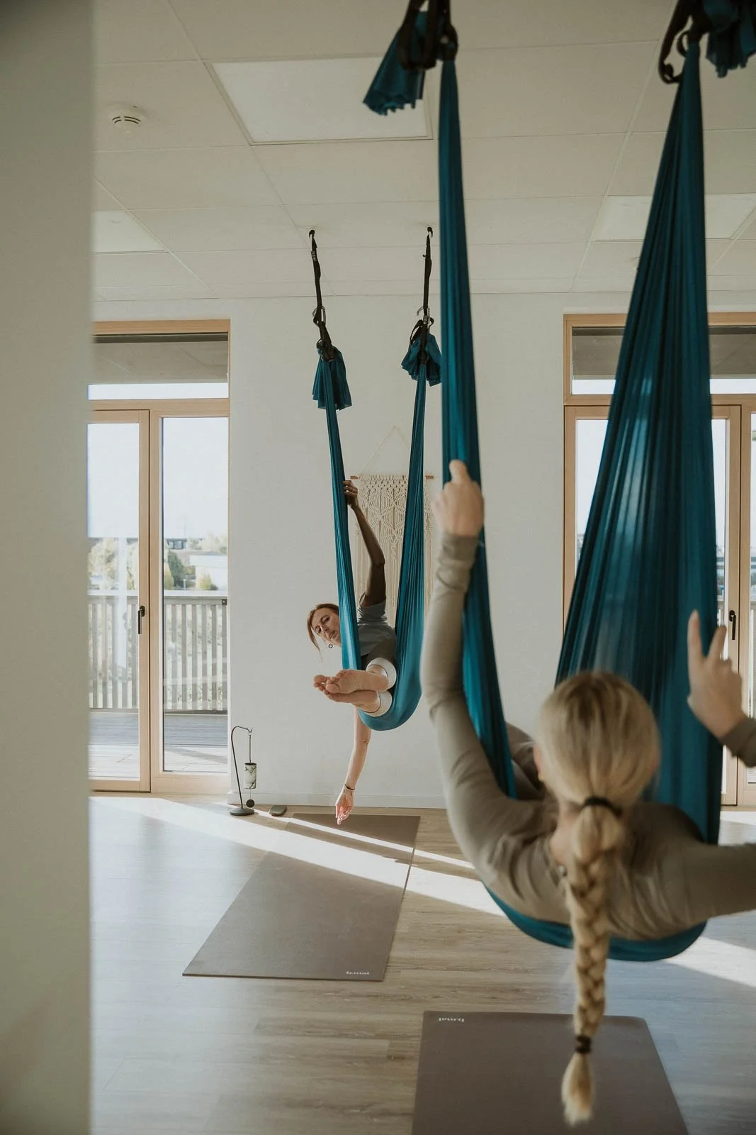 Zwei Frauen im hellen Yogastudio beim Aerial Yoga, schwebend in blauen Tüchern über Yogamatten.