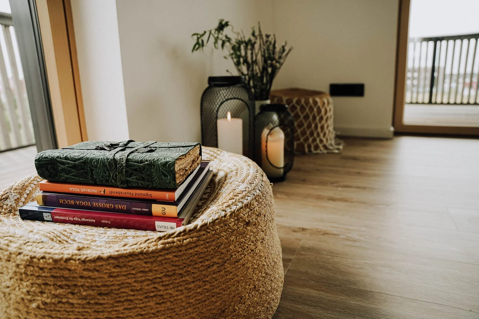 Ein runder, geflochtener Korb mit Büchern und einer zylinderförmigen Kerze auf einem Holztisch, im Hintergrund sind dekorative Laternen mit Kerzen, eine Vase mit Zweigen und ein Sitzkissen auf einem Holzboden.