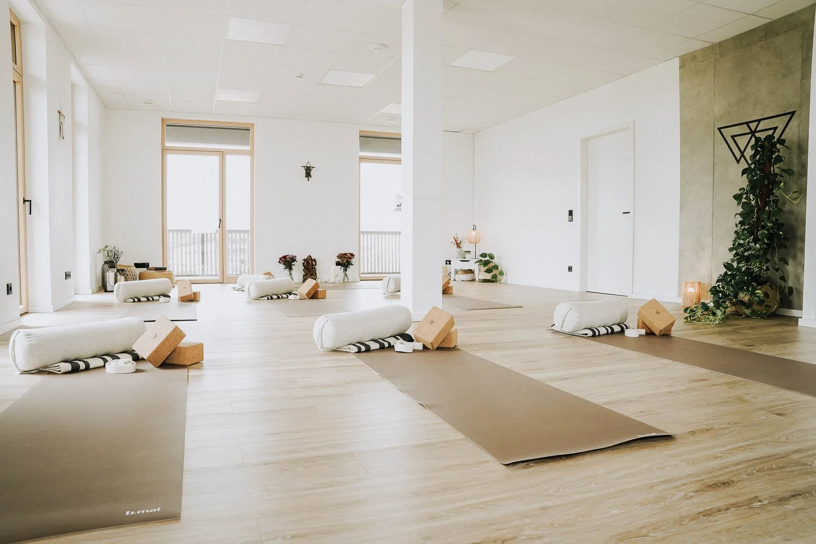 Ein heller, minimalistischer Yoga-Studio-Raum mit Yogamatten, Yogoblöcken und Yogorollen, eingerichtet für eine Yoga-Klasse, mit natürlichem Licht durch große Fenster.