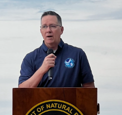 Man with glasses holding microphone at podium with beach and lake in background