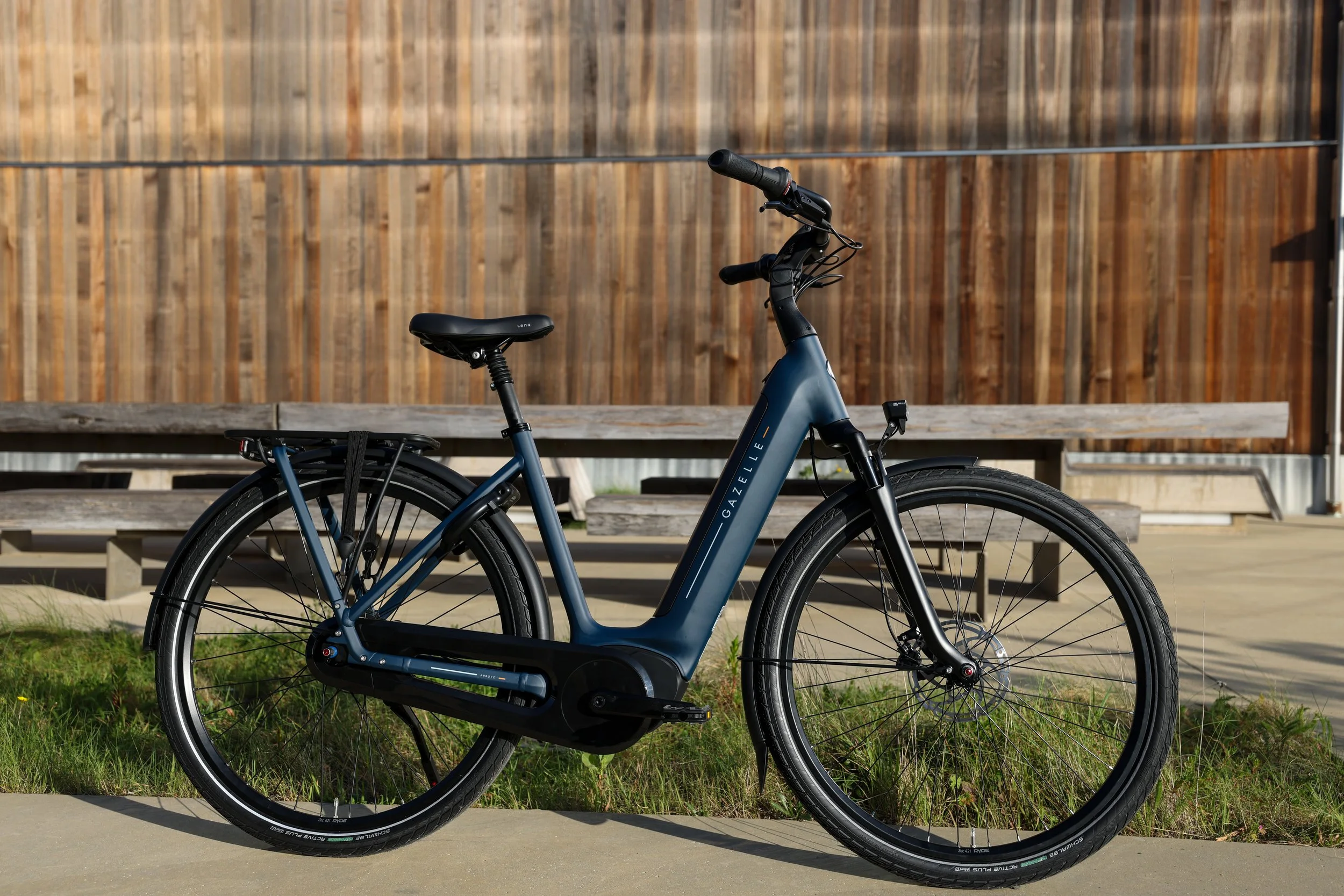 A blue electric bicycle parked on a sidewalk in front of a wooden fence with a bench behind it.