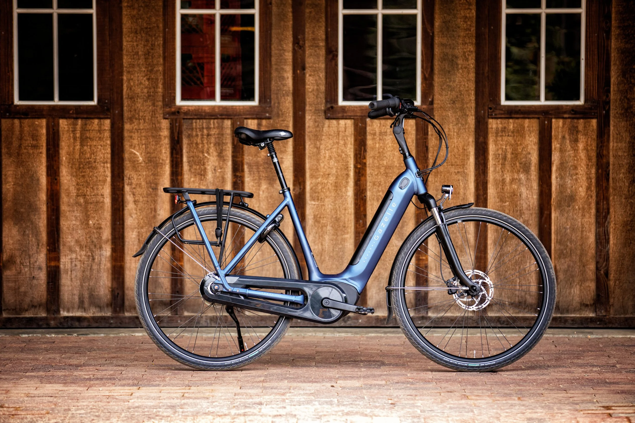 A blue electric bike with a black rear rack parked in front of a wooden wall with three windows.
