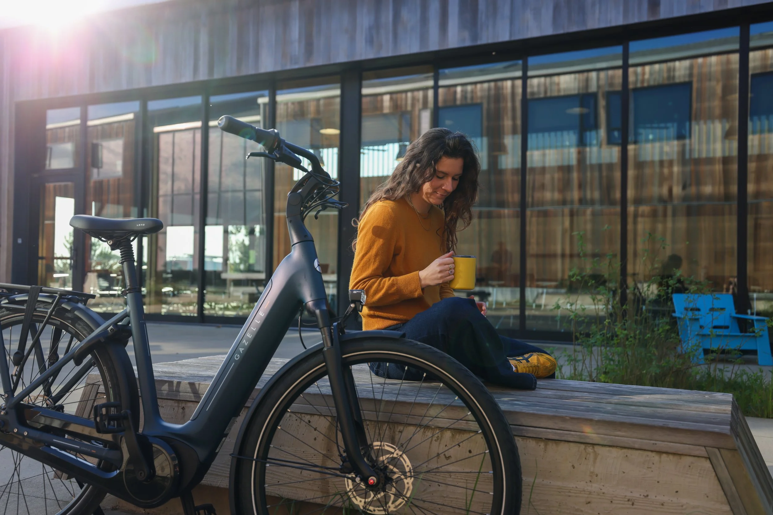 A woman with long dark hair sitting on a bench outside a modern building, holding a yellow mug, with a bicycle nearby.