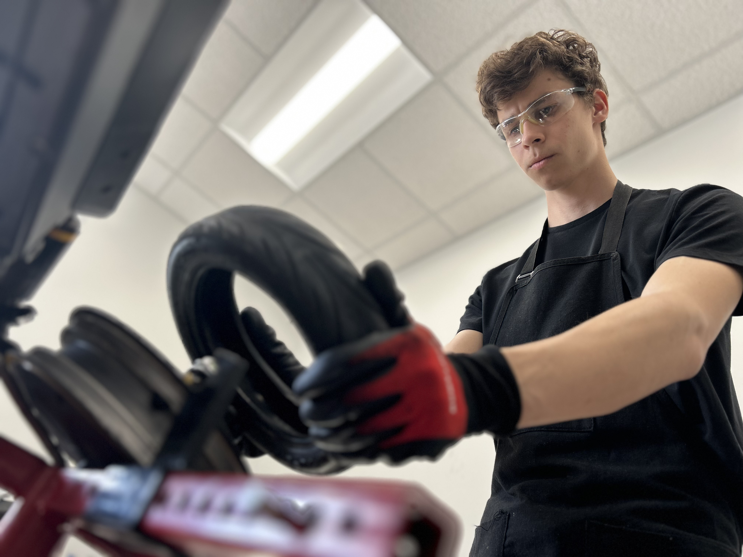 A young man wearing safety glasses and a black apron is working on a bicycle, focusing on the handlebar area in a well-lit room.