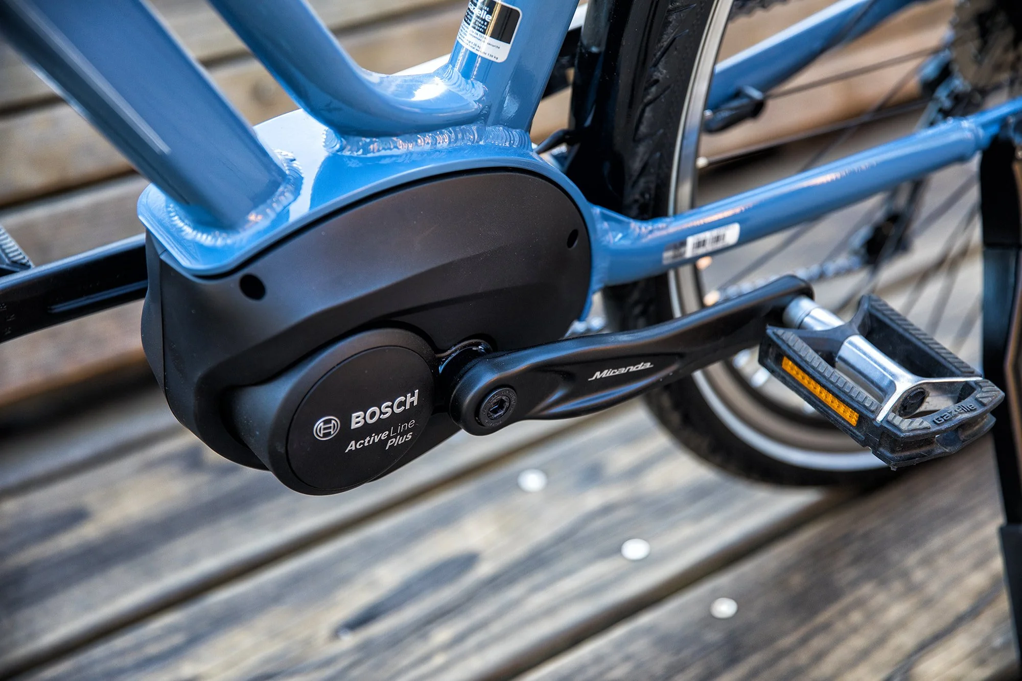Close-up of a blue electric bicycle with a Bosch Active Line Plus motor, pedal, and part of the frame visible, mounted on a wooden surface.