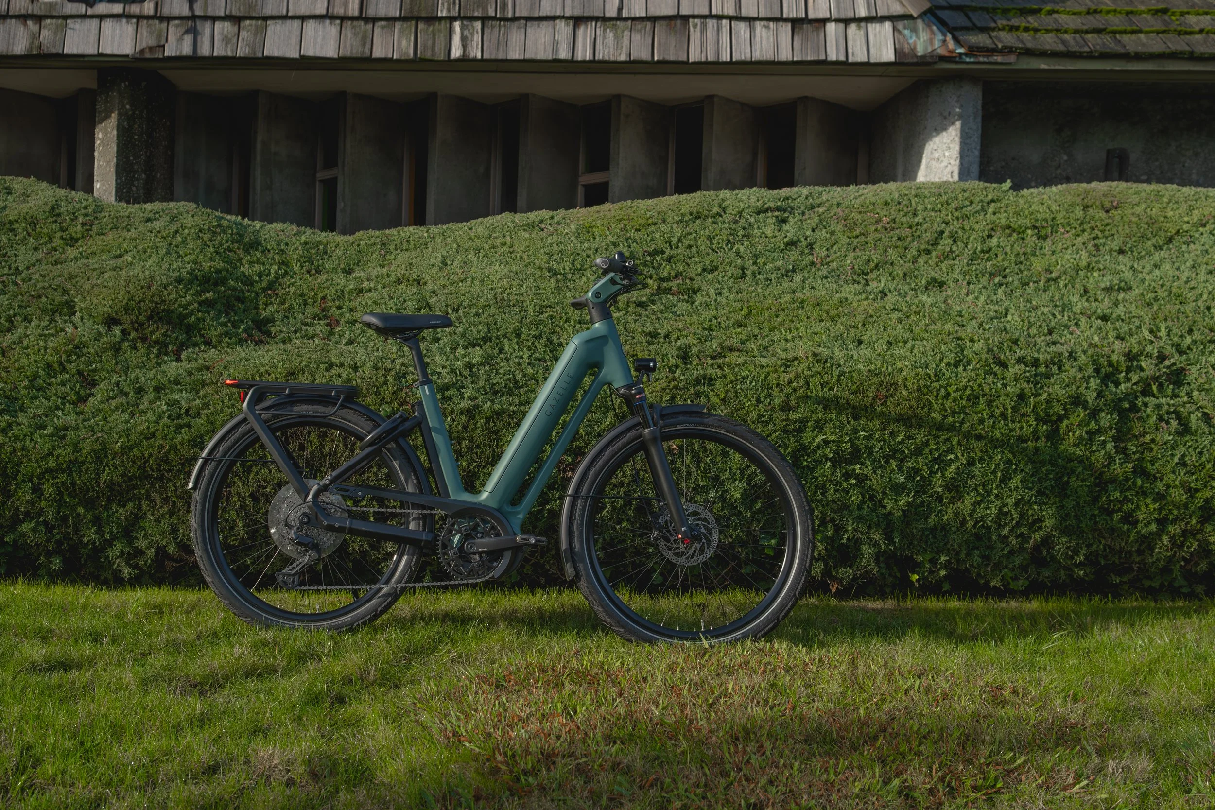 A green electric bicycle parked on a patch of grass in front of dense green bushes. There is a building with a sloped roof and concrete pillars in the background.