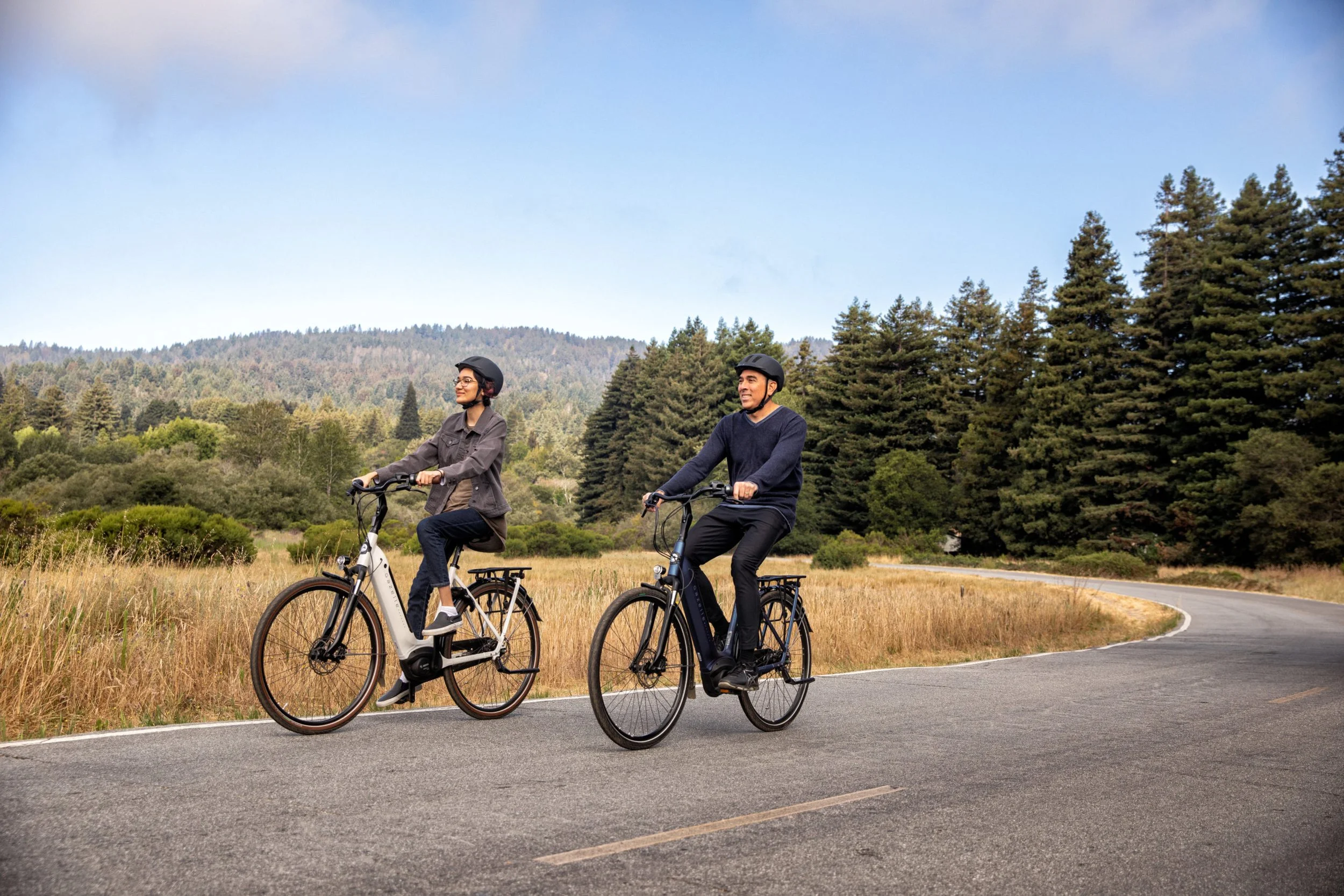 Two people riding electric bicycles on a winding road through a grassy area with trees and hills in the background, wearing helmets.
