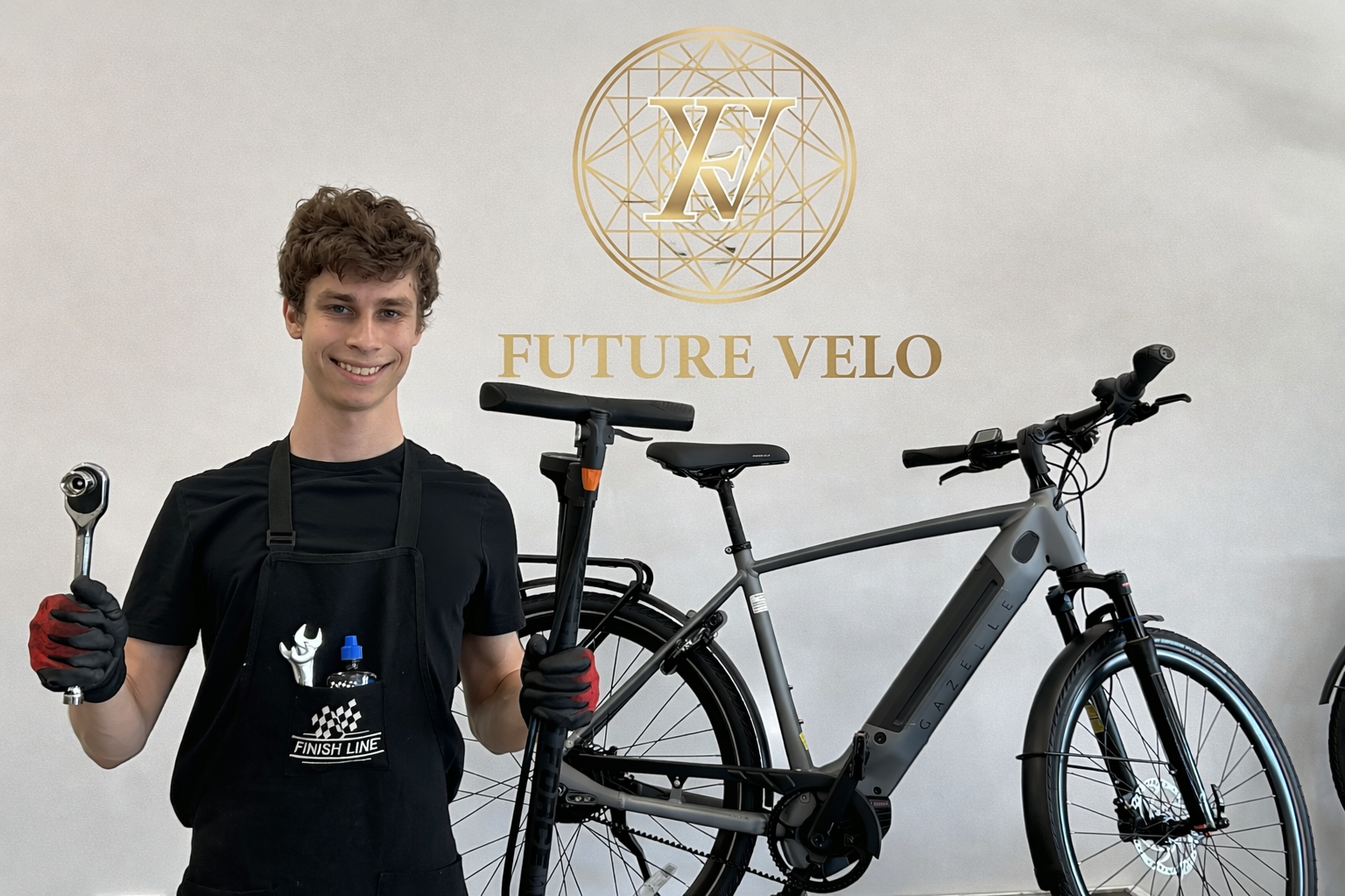A young man wearing a black shirt and gloves holding a wrench and a bike lane marker, standing in front of a wall with a logo and the text "FUTURE VELO" above a bicycle.
