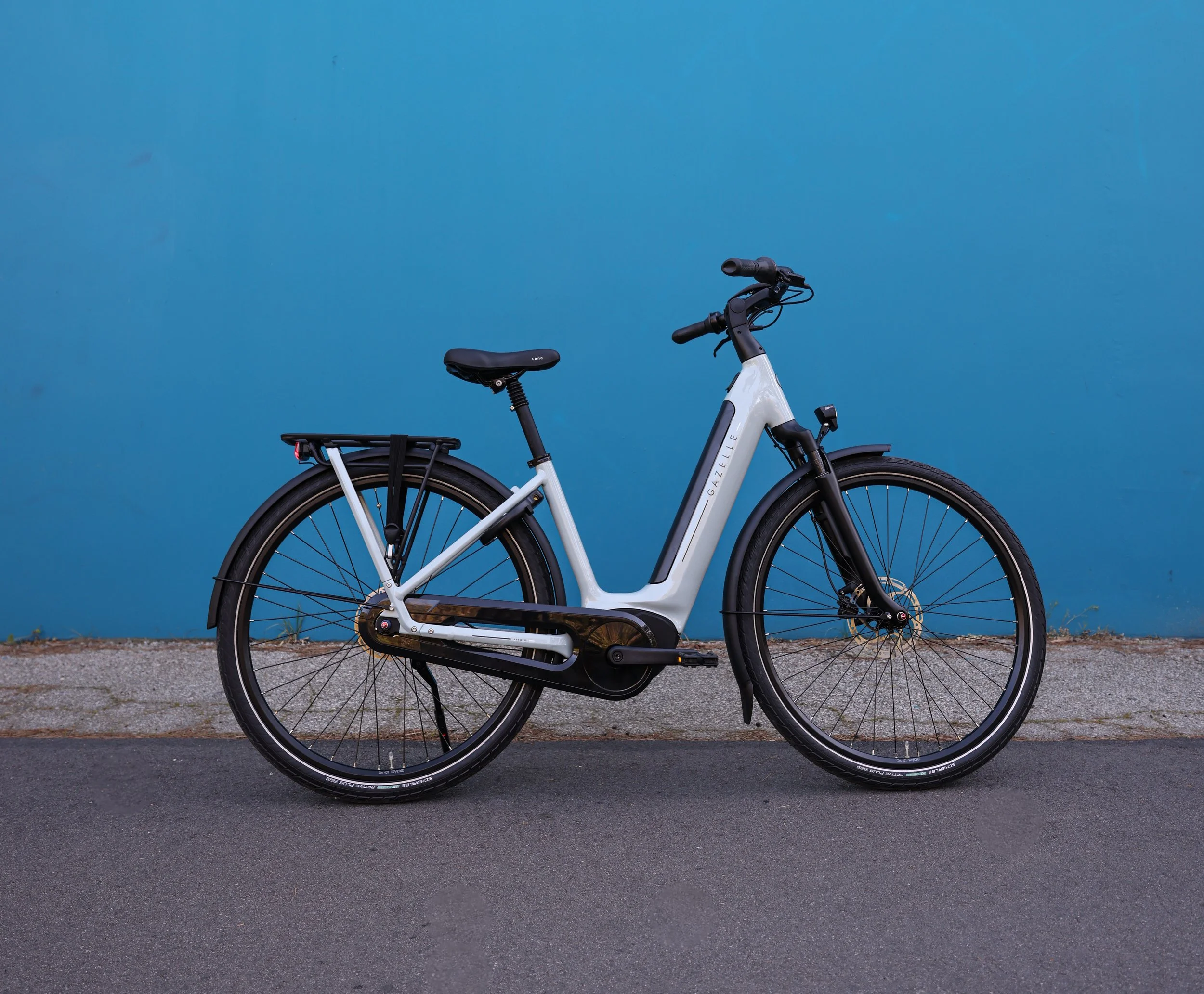 A white e-bike with black accents parked on a paved surface in front of a plain blue wall.