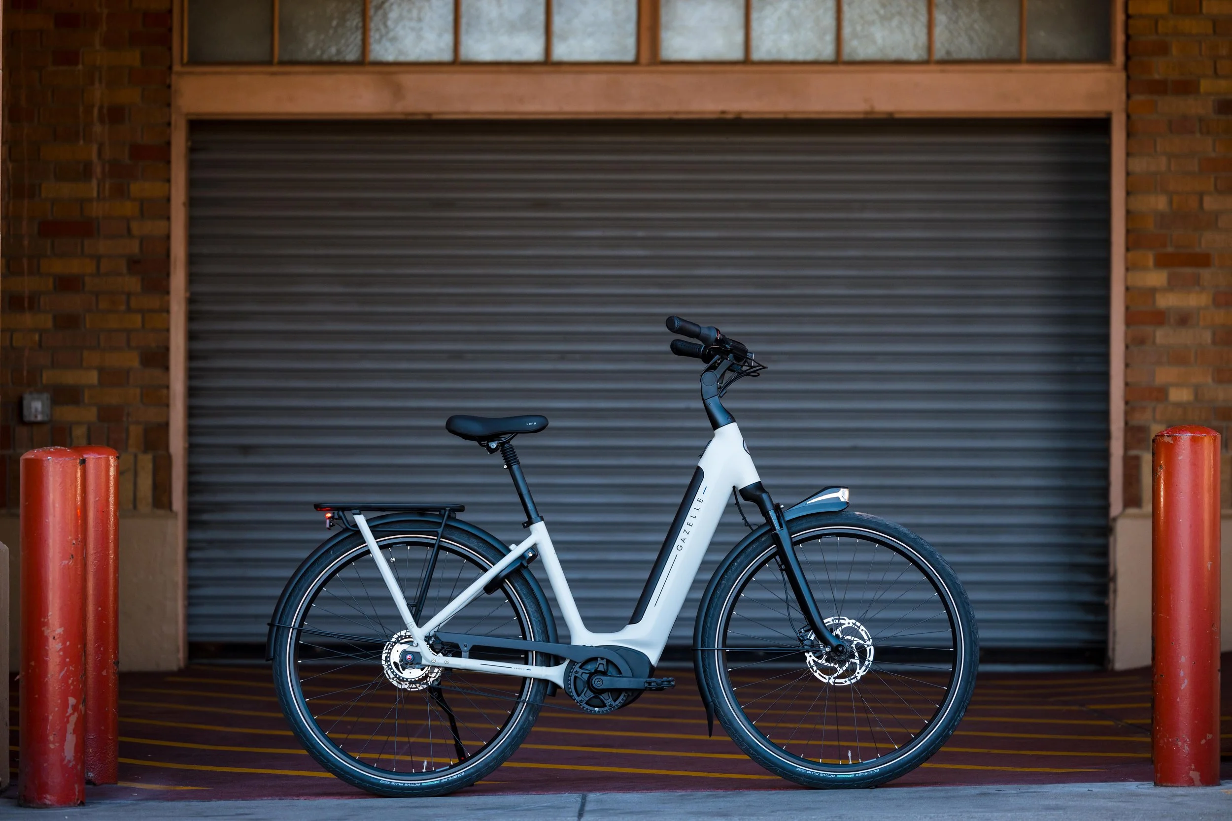 White electronic bicycle parked in front of a closed roll-up garage door, flanked by red bollards, with brick walls on either side.