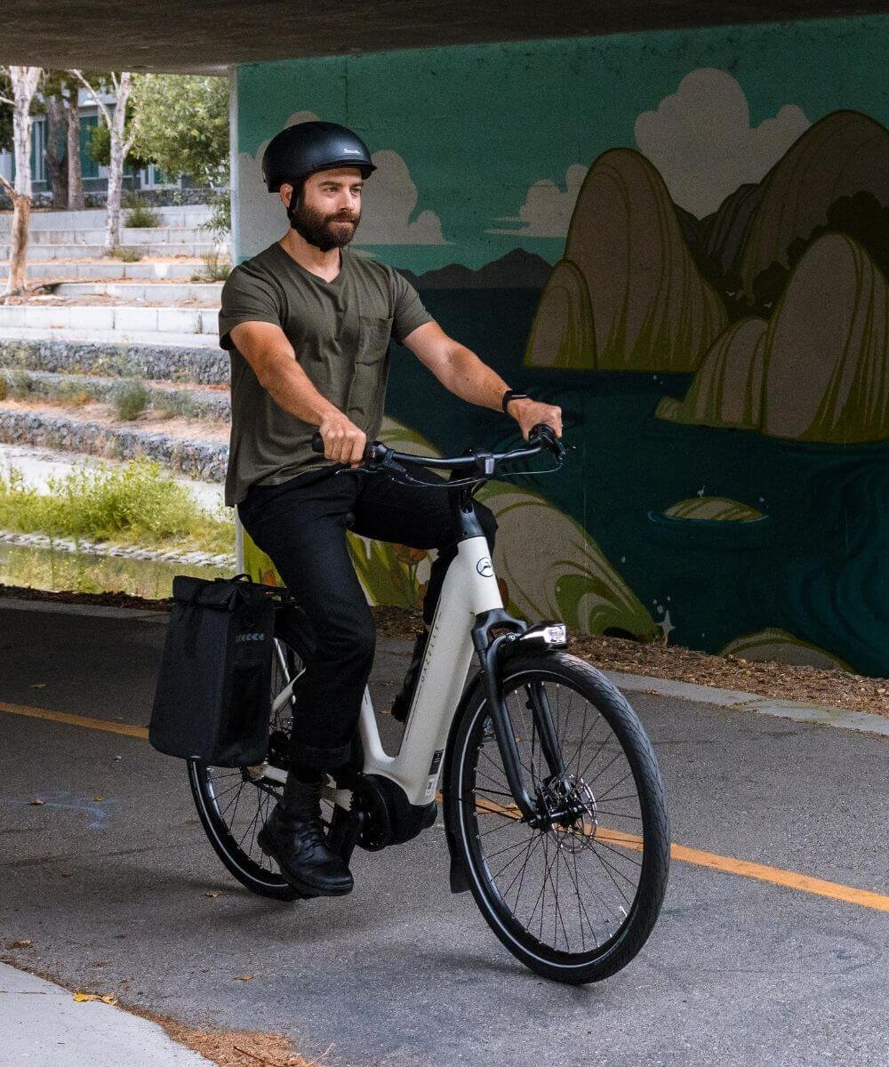 A man wearing a black helmet and casual clothing rides an electric bicycle in an underpass with a painted mountain landscape on the wall behind him.