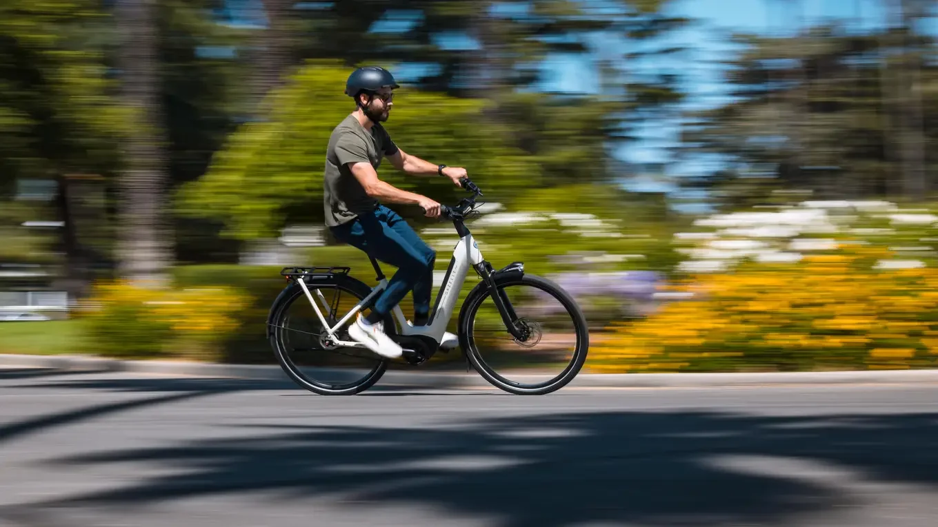 Two people riding bikes on a paved trail surrounded by greenery, both wearing helmets and sunglasses, with the woman in front smiling.