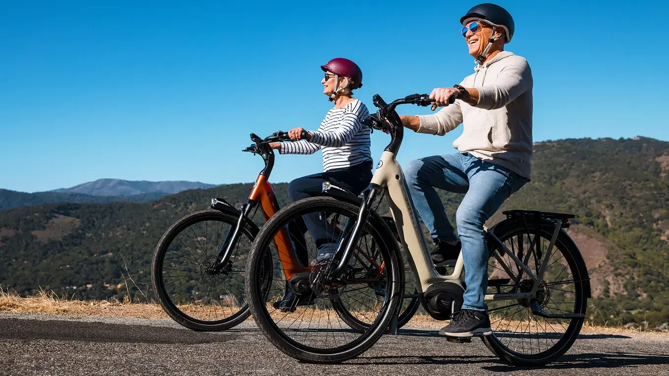 Two older women riding electric bikes on a scenic mountain road, smiling and wearing helmets on a sunny day.