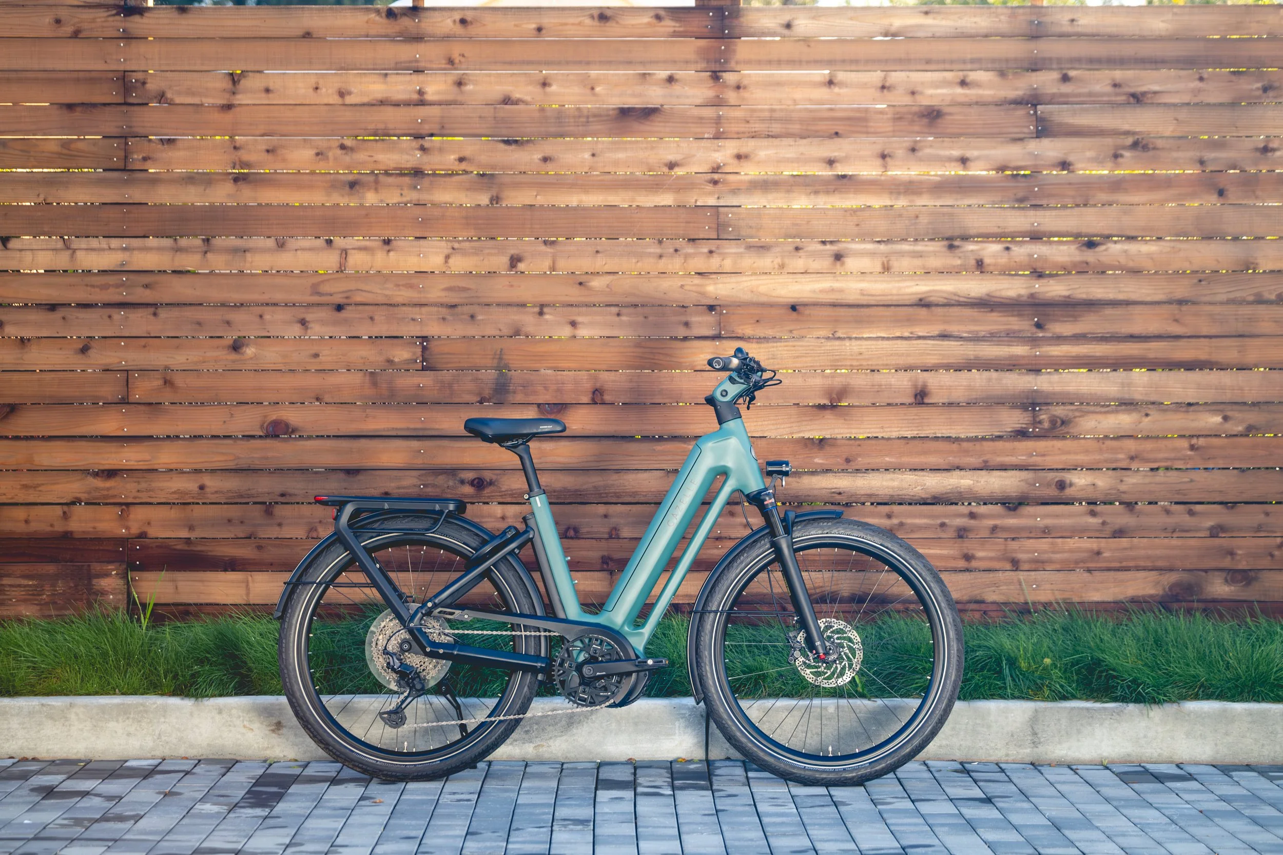 A teal electric bicycle parked on a sidewalk in front of a wooden fence with grass and pavement underneath.