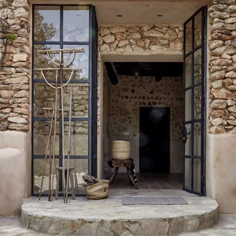 Stone and metal framed open double doors leading into a rustic interior with a stone wall, a small wooden table, a woven basket, and a clay vessel outside.