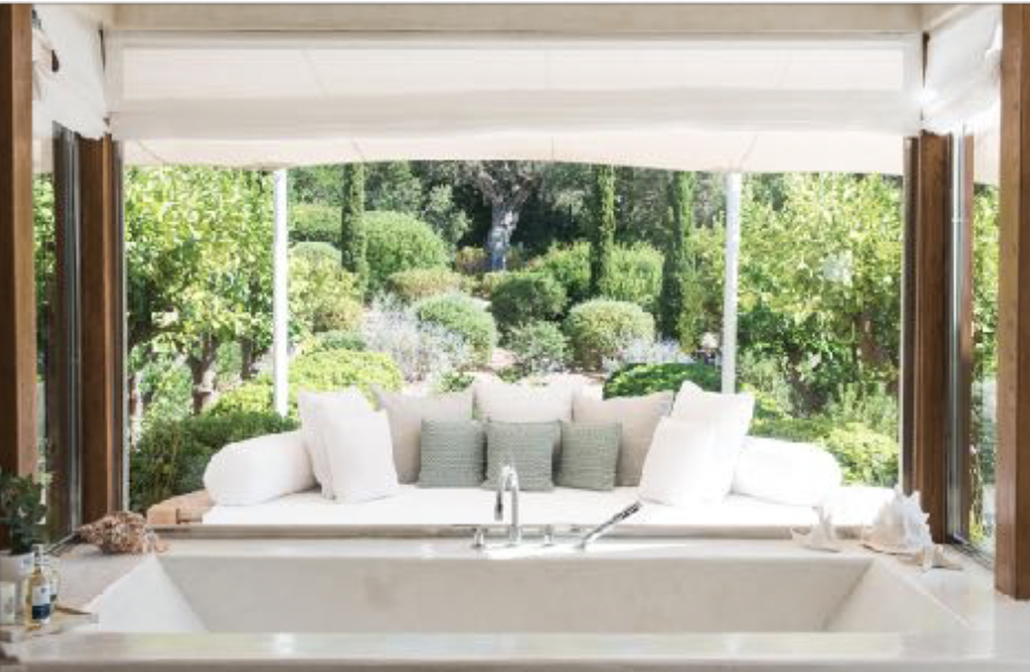 Indoor bathtub facing a large window with a view of lush green garden, cushions on the window seat, and soft lighting.