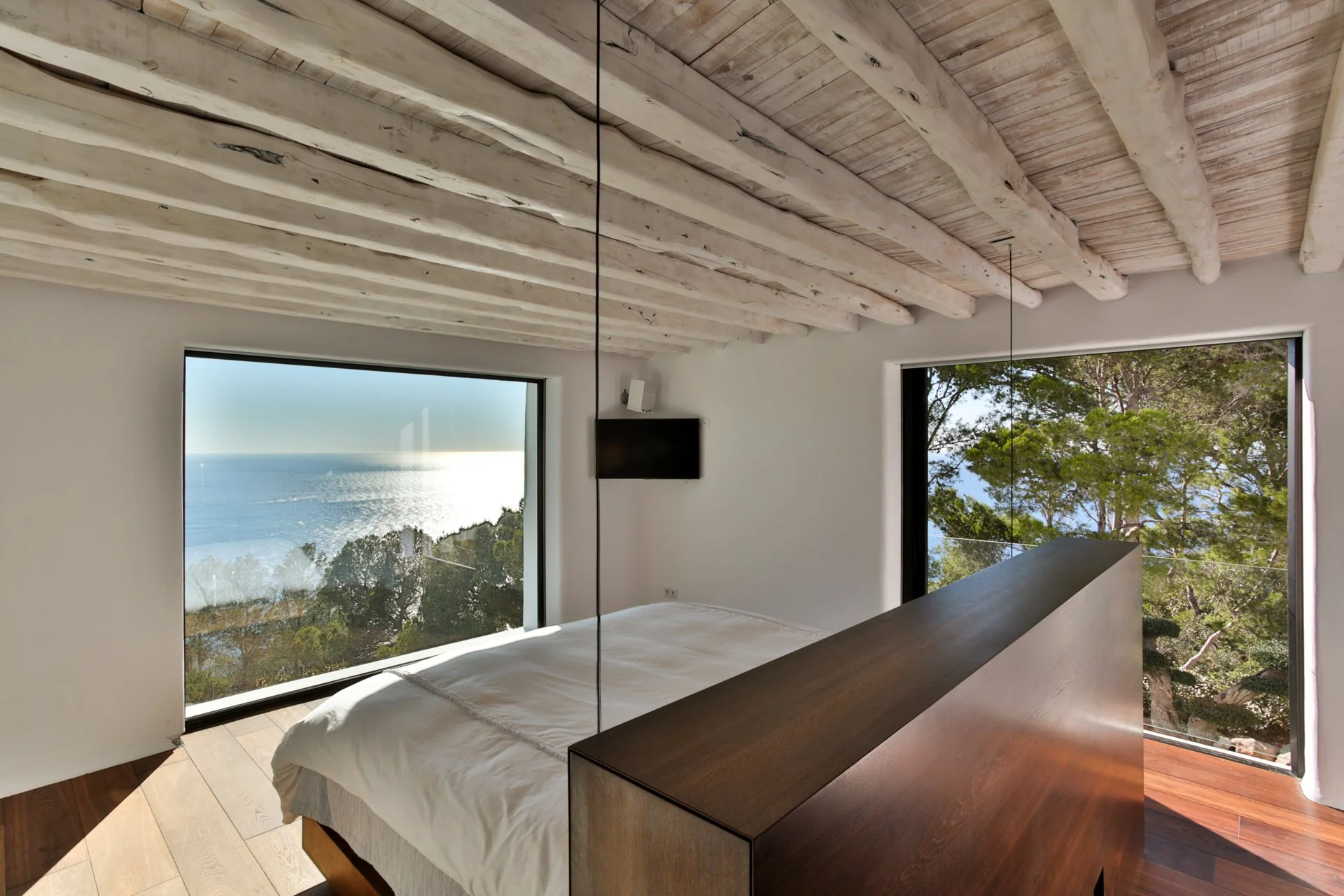 A modern bedroom with large windows showing a view of the ocean and trees. The ceiling has exposed wooden beams painted white, and there is a flat-screen TV mounted on the wall.