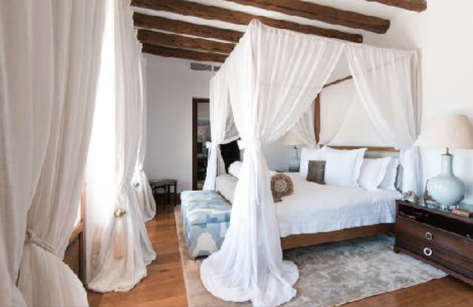 Luxurious bedroom with a four-poster bed, white curtains, and white bedding, wooden beams on the ceiling, and wooden nightstands with decorative vases.