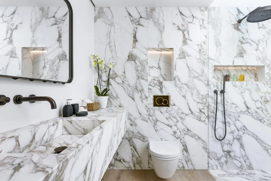 Modern bathroom with marble walls and countertops, a rectangular sink with black fixtures, a mirror, a vase with orchids, a toilet, and a walk-in shower with a black showerhead and built-in shelves.