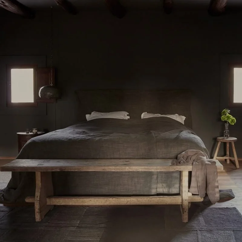 A cozy bedroom with a large bed, dark bedding, and two white pillows. There are two small windows on either side of the bed, with a wooden bench at the foot of the bed, and nightstands with lamps and flowers.