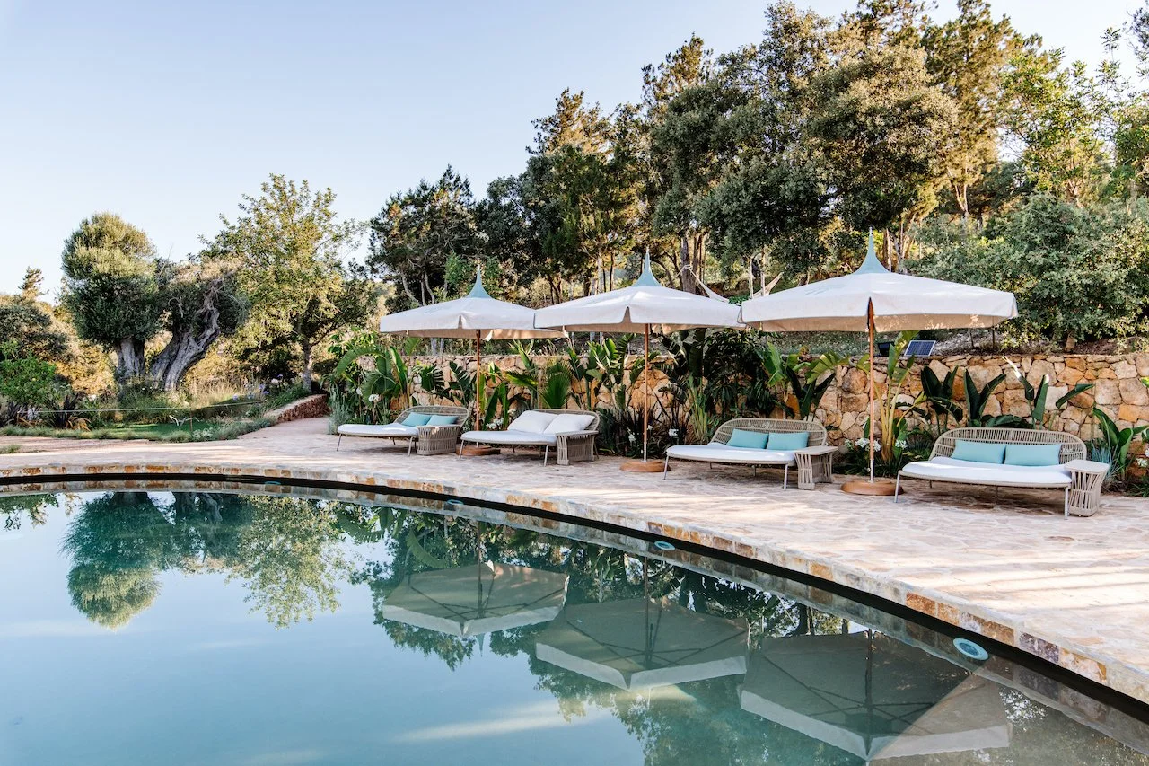 Poolside lounge chairs with umbrellas along a stone patio, surrounded by greenery and trees, reflecting in the water.