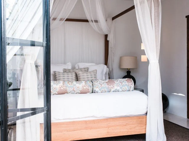 A cozy bedroom with a white four-poster bed with sheer curtains, decorated pillows, a side table with a lamp, and a sliding glass door with curtains.