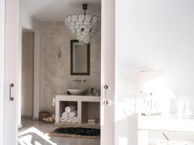 Interior view of a minimalist bathroom with a vanity, bowl sink, oval mirror, rolled towels, a basket, and a decorative chandelier.
