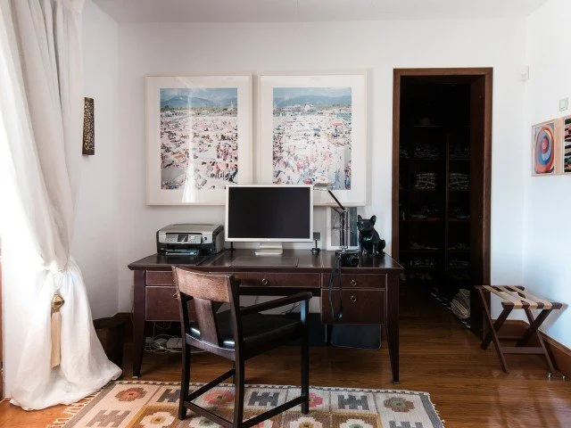 Home office with a wooden desk, computer monitor, printer, and office supplies, a brown chair, framed cityscape artwork on the wall, a dark wood shelving unit, a small bench, and a patterned area rug.
