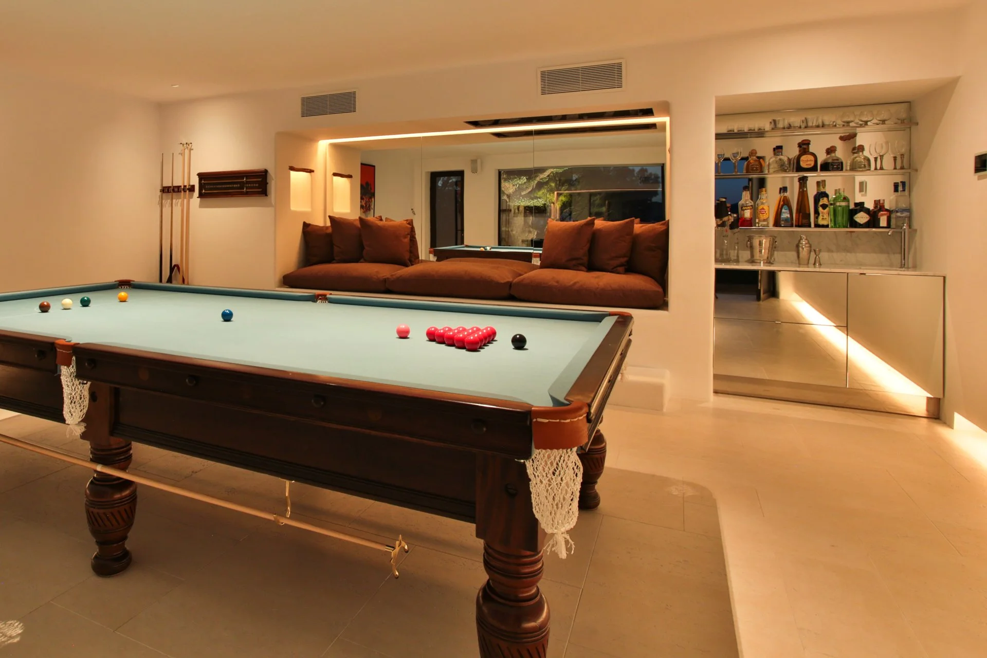 A room with a pool table in the foreground and a built-in bar with bottles and glasses in the background. There is a brown sofa with cushions and a pool table with balls and cues.