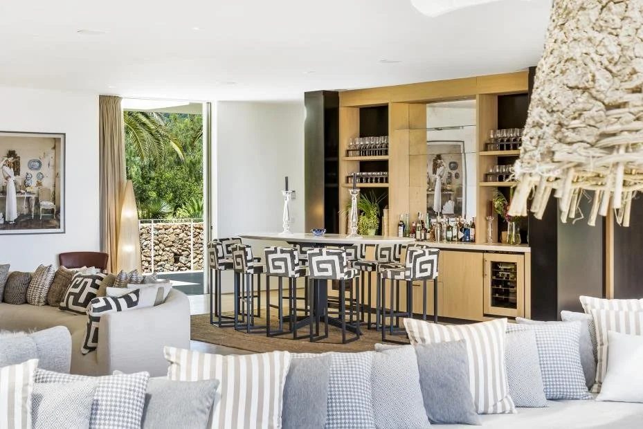 Living room with a bar area featuring patterned barstools, a variety of liquor bottles, glassware, and a large window with curtains, overlooking greenery outside.