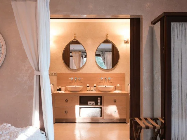 Bathroom with two vessel sinks, oval mirrors above each, wooden vanity, beige walls, and a curtained window on the right.