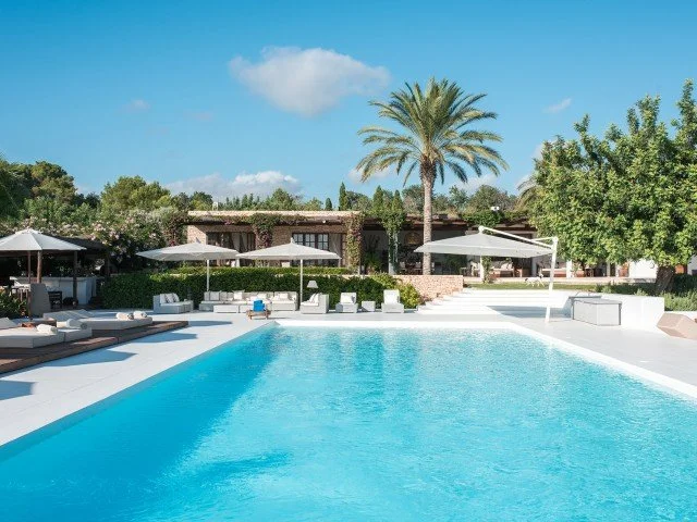 Luxury outdoor swimming pool with white lounge chairs and umbrellas, surrounded by lush greenery and palm trees under a blue sky.
