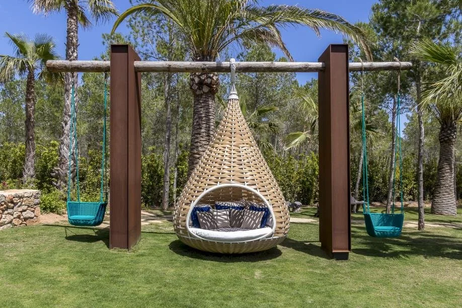 Outdoor swing set with two swings and a hanging wicker egg-shaped chair with cushions, set in a garden with palm trees and green grass.
