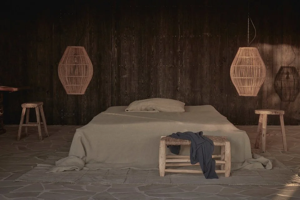 A cozy bedroom with a large bed draped in beige linens, flanked by wooden side tables with woven pendant lights hanging above, and a small bench at the foot of the bed with a dark shirt draped over it. The background features a dark wooden wall and t