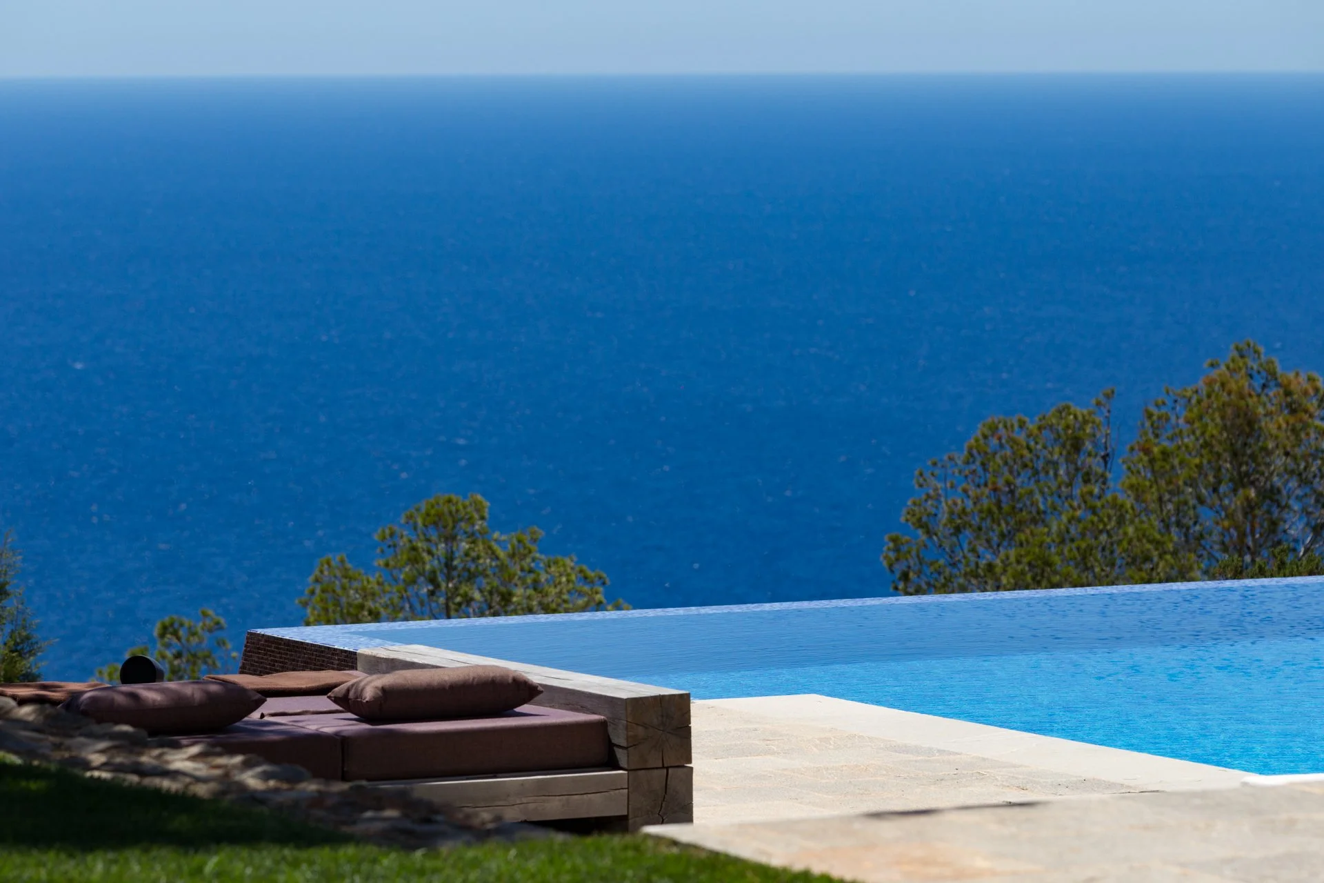 View of an infinity pool overlooking the ocean with lush trees in the background, lounge chairs and cushions on the patio.