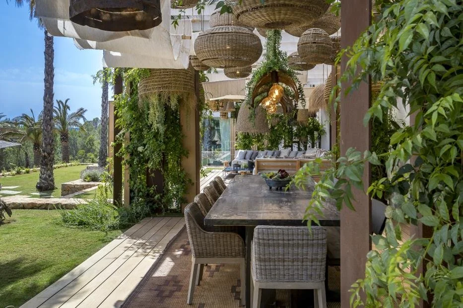 An outdoor dining area with a large wooden table surrounded by wicker chairs. Hanging wicker lamps and potted plants adorn the space, overlooking a lush garden with palm trees and a view of the ocean in the background.