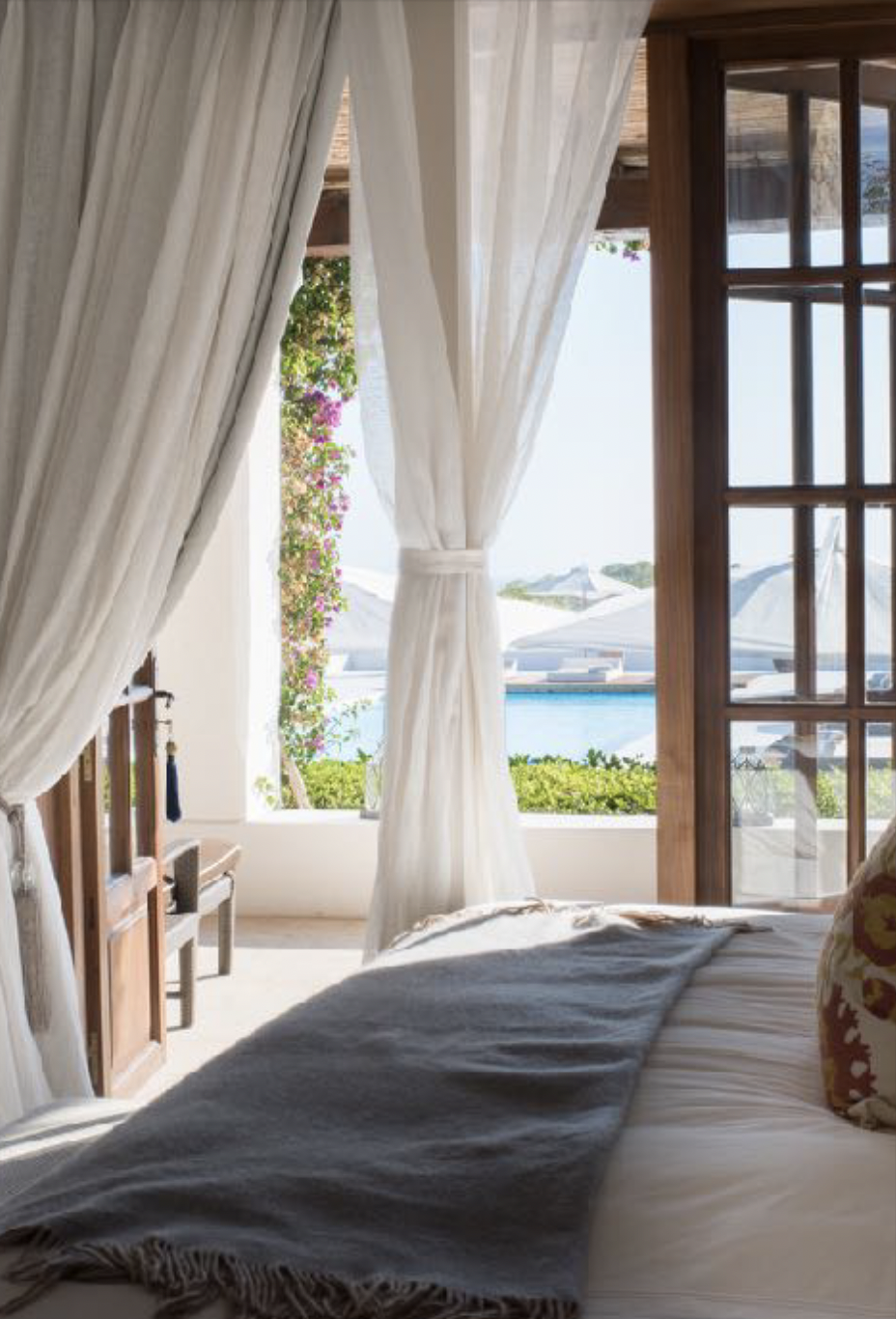 A cozy bedroom with white curtains pulled back, revealing a view of a pool, umbrellas, and greenery outside.