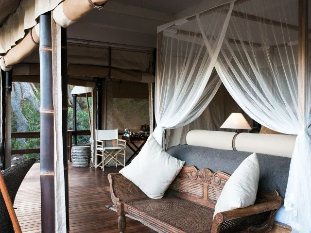 Interior of a luxurious lodge or tent with a canopy bed draped in white curtains, a wooden sofa with white cushions, a desk with a chair, and large windows overlooking greenery.