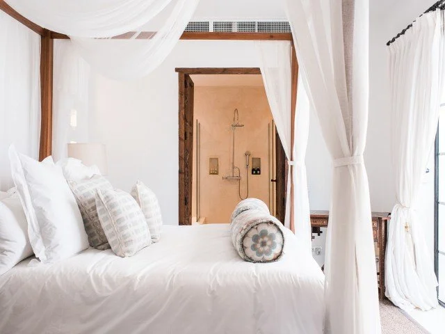 A cozy bedroom with a four-poster bed draped in white curtains, white bedding, a cylindrical decorative pillow, and a view into a small en-suite shower area with beige tiles and a rain shower head.