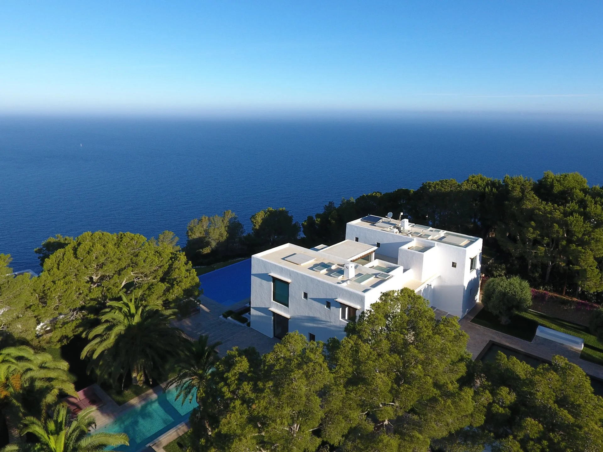 Aerial view of a modern white house surrounded by lush green trees, with the ocean and a clear blue sky in the background.