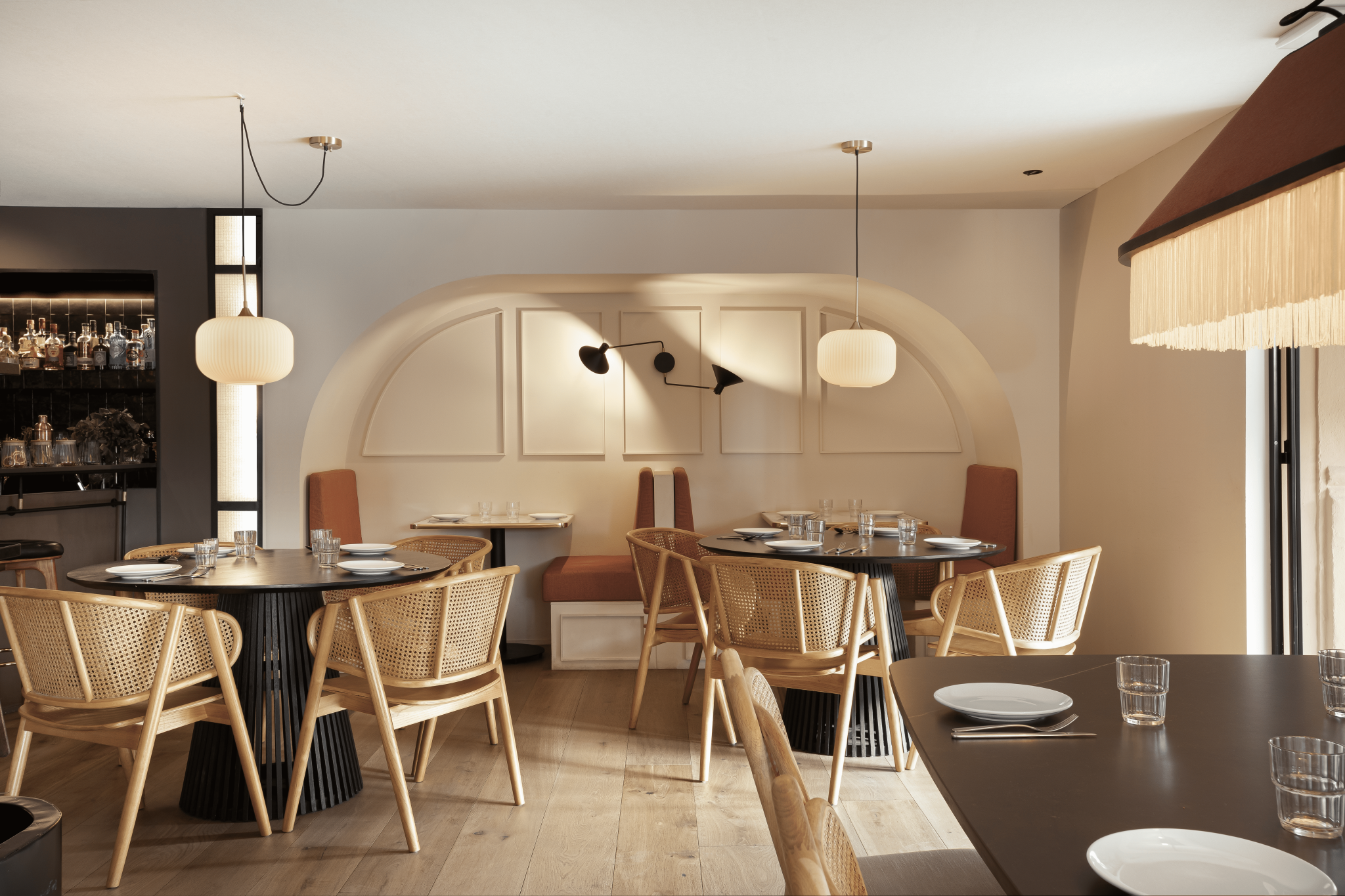 Modern restaurant dining area with round black tables, wicker chairs, and beige booth seating, decorated with white wall panels and contemporary lighting fixtures.