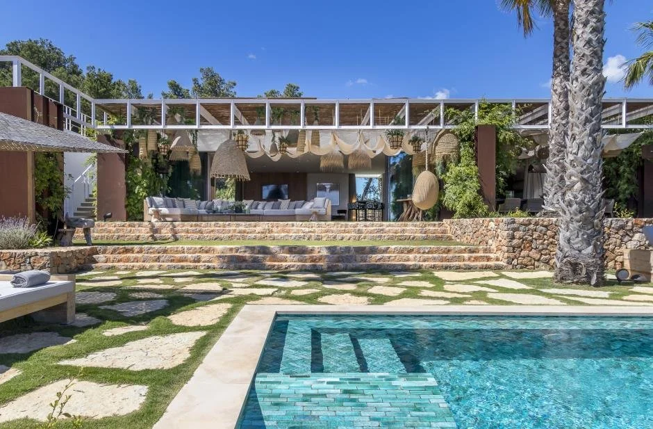 Modern backyard with pool, stone steps, outdoor sofa, hanging woven lamps, and lush greenery under a bright blue sky.