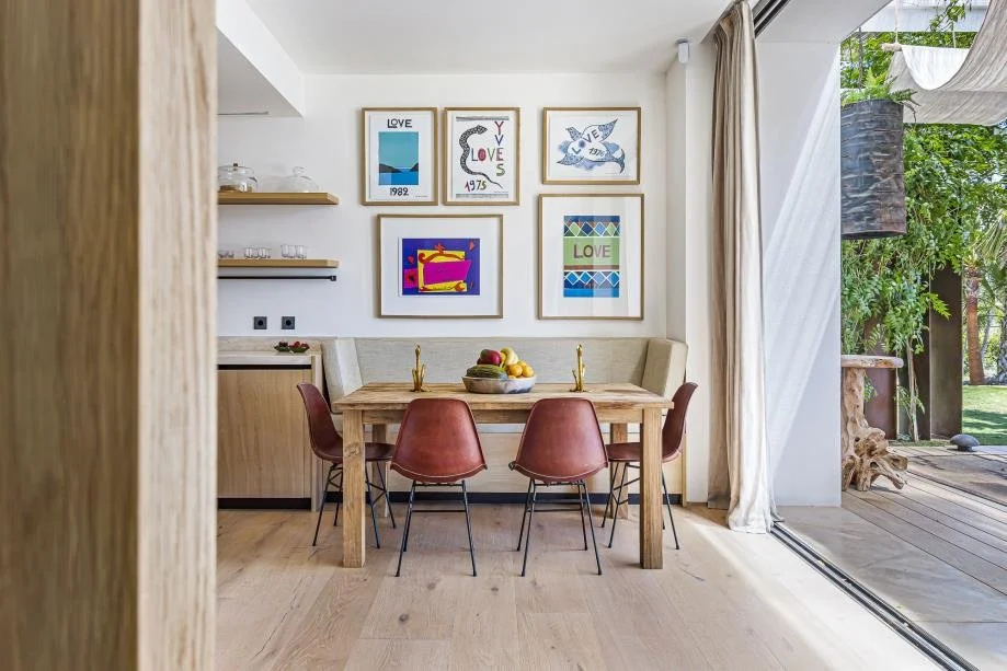 Dining area with a wooden table, four brown chairs, a fruit bowl, and a framed art gallery on the white wall, adjacent to an open sliding door leading to a patio with greenery.