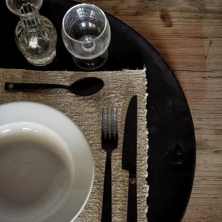 Table setting with a white bowl, black fork, black knife, black spoon, a glass, and a small bottle on a woven placemat, on a dark round table with a wooden floor background.