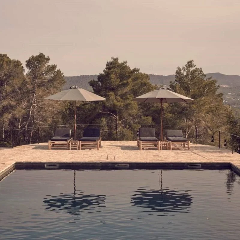 Poolside scene with four lounge chairs and two large umbrellas on a deck with a scenic view of trees and mountains in the background.