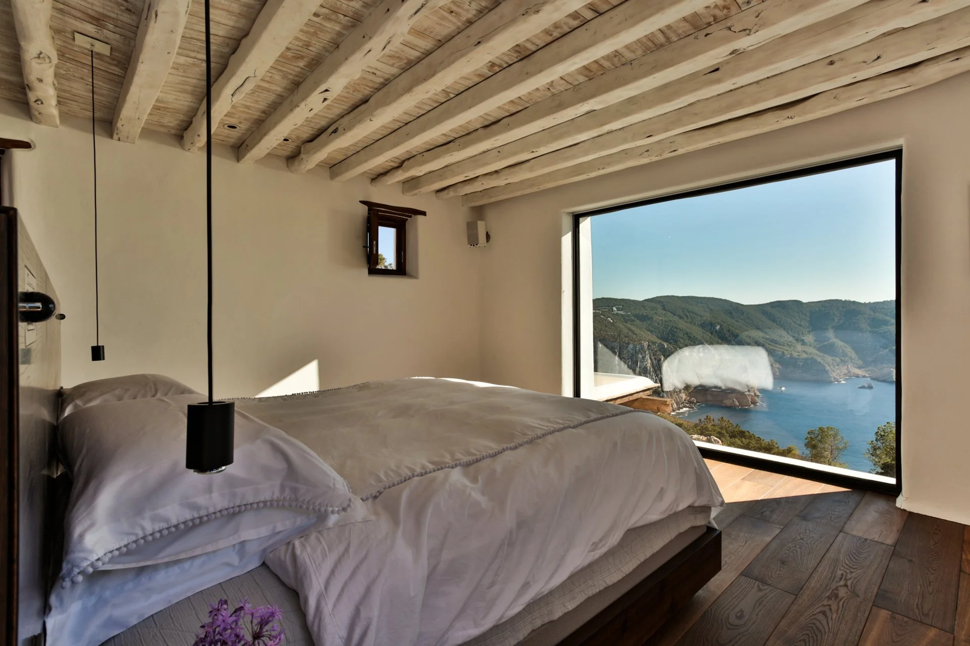 A bedroom with a large window showing a coastal landscape with hills and the ocean, a bed with white bedding, wooden ceiling beams, and small wall-mounted lamps.