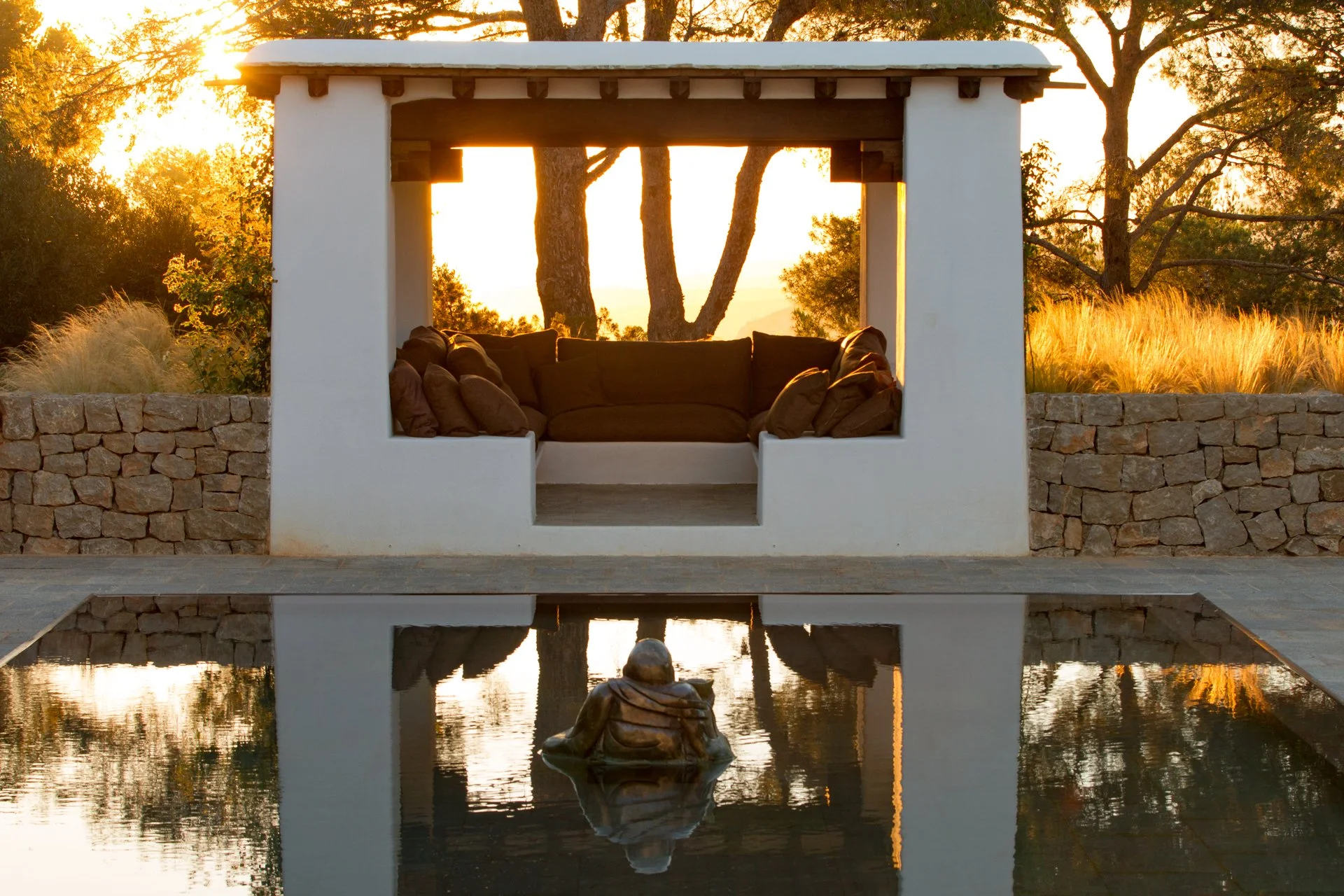 A white outdoor structure with an open center filled with brown cushions, overlooking a pond with a reflected statue of a sitting person, trees, and a sunset in the background.