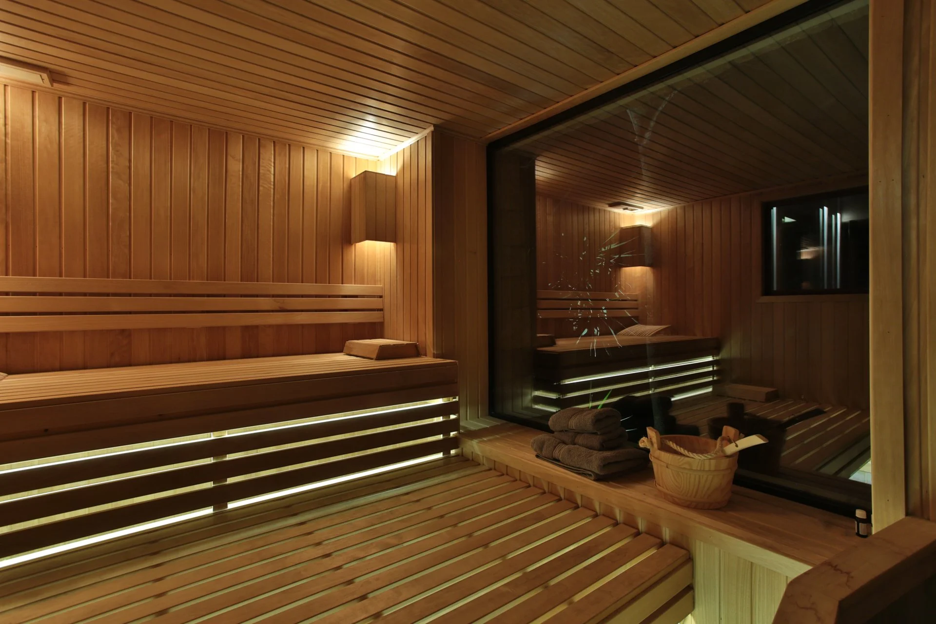 Cozy indoor sauna room with wooden paneling, benches, stacks of towels, and a glass window showing the interior reflection.