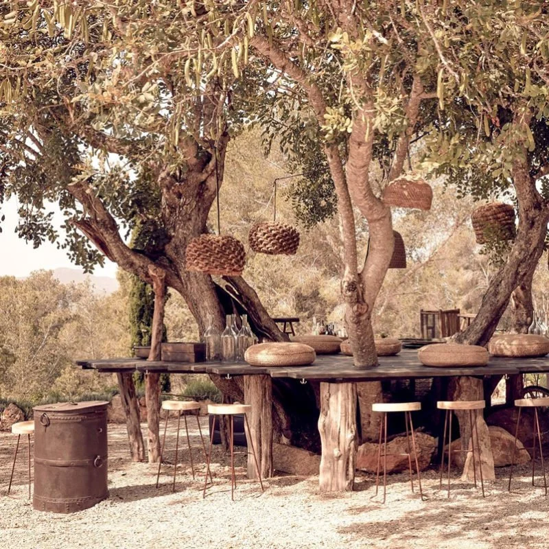 Outdoor seating area under large trees with woven hanging lamps and wooden stools, in a rustic, natural setting.