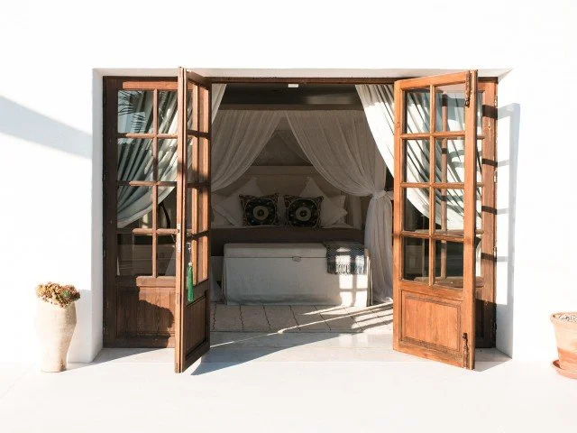 Open wooden French doors revealing a bedroom with a bed and sheer curtains.